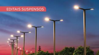 Imagem mostra vários postes de iluminação em perspectiva. O céu ao fundo vai em degradê do azul para o roxo. No alto, à esquerda, há a inscrição Editais Suspensos em branco sobre uma tarja vermelha