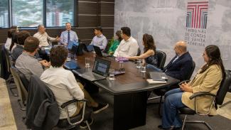 Imagem mostra 14 pessoas sentadas ao redor de uma mesa marrom, onde há computares sobre ela