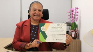 Foto da diretora da Ouvidoria do TCE/SC, Walkiria Machado Rodrigues Macie, em sua sala de trabalho. Ela é uma mulher negra, usa um paletó vermelho sobre uma blusa marrom com bolinhas azuis. Ela segura o certificado que recebeu.