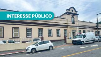 Banner horizontal com foto da fachada do Hospital da Polícia Militar Lara Ribas. É uma construção com arquitetura antiga, com paredes bege, detalhes em marrom, diversas janelas e uma torre central com janela em arco. Em frente ao edifício, na rua, há um carro branco estacionado e uma ambulância. Sobre a foto, em tarja verde-água, o texto "Interesse público".