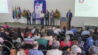 Foto horizontal que mostra, ao fundo, o conselheiro Herneus De Nadal falando ao púlpito. Ao lado dele, há cinco pessoas, entre elas o conselheiro substituto Gerson Sicca. À frente do presidente, há diversos servidores sentados em cadeiras, um ao lado do outro. 