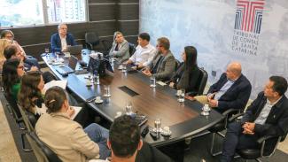 Banner horizontal com foto de reunião realizada no TCE/SC. Há várias pessoas sentadas ao redor de uma mesa, enquanto conversam. Na ponta, o conselheiro substituto Gerson dos Santos Sicca.  