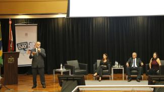 Imagem mostra quatro pessoas em um palco com fundo marrom e um banner, à esquerda, onde se destaca a expressão Ouvidoria Day. Em pé, à esquerda, o diretor da Ouvidoria do TCE/SC fala ao microfone. Sentados, da esquerda para a direita, estão  ouvidora-geral do Estado, Marina Rebelo; o ouvidor do MPSC, procurador Paulo Cezar Ramos de Oliveira; e a auditora federal de Finanças e Controle da CGU, Rosemary Zucarelli.