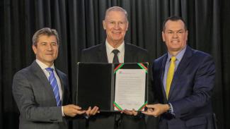 Banner horizontal com foto dos conselheiros José Nei Ascari, Herneus De Nadal e Adircélio de Moraes Ferreira Júnior no ato de posse. Eles estão em pé, um ao lado do outro, sorrindo, no palco do auditório do TCE/SC. Ambos seguram e mostram para a foto um documento assinado, dentro de uma prancheta. Atrás deles, as demais autoridades que compõem a bancada batem palmas. 