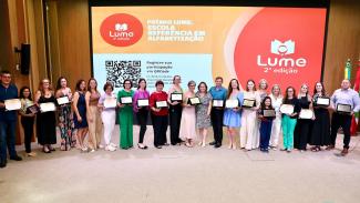 Foto que mostra representantes de todas as escolas premiadas no palco do auditório do evento. Há 23 pessoas em pé, uma ao lado da outra. A maioria segura os certificados e mostram-nos para a foto. Atrás, no telão, é projetado o logo do prêmio Lume, em tons de laranja. 