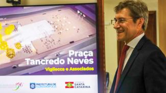 Banner horizontal com foto do conselheiro José Nei Ascari sorrindo, à direita. Ele é um homem branco, de cabelos grisalhos, que veste terno e gravata. Ao lado esquerdo, uma televisão mostra o projeto da Praça Tancredo Neves