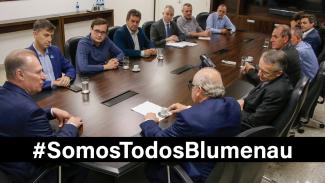 Banner horizontal com a foto dos participantes da audiência. Estão sentados em volta de uma mesa, 13 homens, entre eles, o presidente do TCE/SC, conselheiro Herneus De Nadal, que está na cabeceira. Sobre a imagem, o texto “#SomosTodosBlumenau”, em fonte branca e sobre uma tarja preta. 