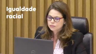 Banner com a foto da conselheira substituta Sabrina Nunes Iocken, uma mulher branca, usa óculos, cabelos castanhos. Ela está sentada em uma cadeira preta. À sua frente, um microfone e parte da tela de um computador. À esquerda da imagem, o texto "Igualdade Racial", em fonte branca.