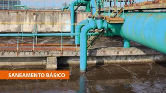 Foto de uma estação de tratamento de esgoto com canos e tubulações azuis. No canto inferior esquerdo, sobre tarja laranja, o texto "Saneamento básico".