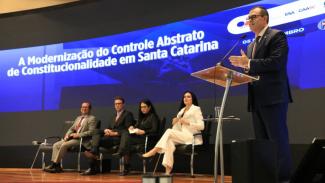 A imagem traz cinco pessoas em um palco, durante uma palestra, sendo que quatro estão sentadas a partir da esquerda para direita: dois homes e duas mulheres. De pé, no canto direito da foto, um homem, conselheiro do Tribunal de Contas de Santa Catarina Adircélio de Moraes Ferreira Júnior, faz uma apresentação em frente a um púlpito. Ele veste terno azul-escuro e está com as duas mães estendidas a frente.