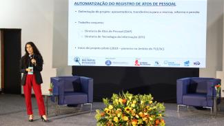 Imagem mostra uma mulher que veste calça vermelha e casaco preto em um palco, do lado esquerdo de um painel onde estão projetadas algumas frases.