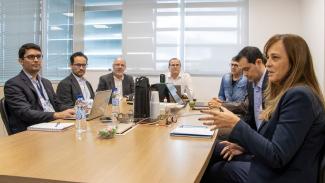 Foto de um grupo de pessoas sentadas ao redor de uma mesa retangular. À direita, uma mulher de cabelos compridos e castanhos. Os outros seis são homens.