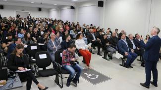 Foto do auditório onde se realizava o evento. No canto direito da imagem, está o diretor-geral de Controle Externo do TCE/SC, Sideny Tavares Jr, um homem branco, cabelos claros e veste um terno azul. Na plateia, homens e mulheres assistem a palestra. 