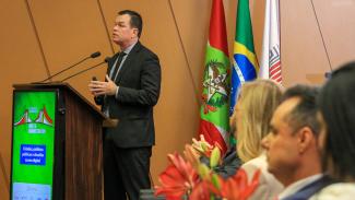 Imagem mostra do lado esquerdo um homem falando em um microfone. Ele veste um traje cinza escuto e está atrás de um púlpito.