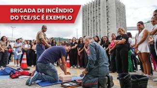 Imagem que mostra simulação de emergência em frente ao Fórum Rid Silva. Há várias pessoas olhando os brigadistas simulando uma massagem cardíaca. Atrás, é possível ver o prédio do Tribunal de Justiça. Sobre a foto, em retângulo bordô, o texto “Brigada de incêndio do TCE/SC é exemplo”.