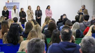 Imagem mostraum grupo de pessoas em um palco, com público em primeiro plano. À direita, há um homem falando em um microfone, atrás de um púlpito. À esquerda aparecem seis mulheres