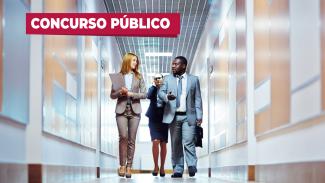 Foto de um corredor, tirada do chão. Três pessoas caminham para frente em direção à câmera: uma mulher loira segurando uma pasta, um homem negro segurando uma pasta, que conversam entre si, e uma mulher morena, de óculos, ao fundo. Na frente da imagem, acima e à esquerda, uma tarja vermelha com o título "Concurso público"