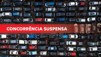 Banner horizontal com foto de grande quantidade de carros estacionados lado a lado, vistos de cima, formando várias fileiras apertadas. Os veículos têm diferentes cores, como preto, branco, vermelho, azul e cinza, e ocupam praticamente todo o espaço da imagem. Sobre a foto, uma tarja vermelha com o texto "Concorrência suspensa".