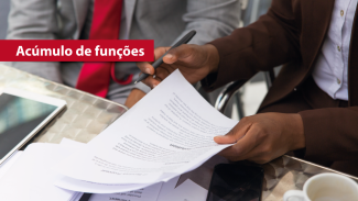 Banner horizontal com imagem de ambiente de trabalho corporativo. Ao centro, mãos de uma pessoa seguram e analisam documentos impressos e seguram uma caneta, sugerindo revisão ou assinatura de papéis. Sobre a mesa, há folhas espalhadas, um celular e um tablet parcialmente visível. No canto superior esquerdo, um destaque em faixa vermelha traz o texto “Acúmulo de funções”.