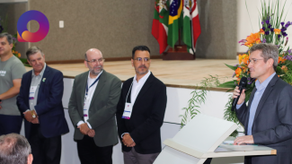 A foto mostra cinco pessoas perfiladas. Em primeiro plano há um homem de casaco escuro falando ao microfone.