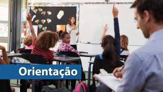 Foto de uma sala de aula em que aparecem alguns alunos sentados nas carteiras e a professora ao fundo, à frente do quadro negro. Em primeiro plano, um homem segura uma prancheta e escreve no papel. No canto inferior direito uma tarja azul com o título “Orientação” em letras brancas. 