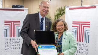 Banner horizontal com foto do presidente do TCE/SC, conselheiro Herneus De Nadal, ao lado da secretária nacional de Integridade, Patrícia Alvares de Azevedo Oliveira. Eles seguram o selo de apoiador institucional do Programa Time Brasil, entregue ao conselheiro Herneus. Ambos sorriem e estão em pé, em frente a dois banners com logo do TCE/SC.