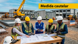 Banner horizontal com foto de uma obra. Em primeiro plano, cinco profissionais usando capacetes e equipamentos de segurança analisam plantas e documentos sobre uma mesa de trabalho. Ao fundo, há estruturas modulares em construção, máquinas pesadas e um guindaste em operação. No canto superior direito, uma faixa amarela destaca o texto “Medida cautelar”.   