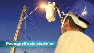 Banner horizontal com foto, vista de baixo, de um homem, com capacete de segurança, observando outro trabalhador que mexe na luz de um poste, suspenso em um equipamento que o ergue. Sobre a imagem, no canto inferior esquerdo, o título “Revogação de cautelar”, em tarja azul. 