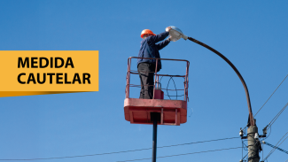 Banner horizontal com foto de um trabalhador usando capacete laranja e uniforme azul, em uma plataforma elevatória vermelha, realizando manutenção em um poste de iluminação pública contra um céu azul. No lado esquerdo, há um retângulo amarelo com o texto "Medida cautelar".