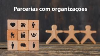 Banner horizontal com imagem ilustrativa de um cubo de madeira com ícones que representam áreas ou temas sociais, como coração, casa, acessibilidade, família, inclusão, diálogo e pessoas. À direita, aparecem três figuras de madeira estilizadas, de mãos dadas. Sobre a foto, na parte superior, lê-se "Parcerias com organizações". 