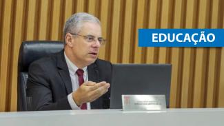 Foto do conselheiro substituto Gerson Sicca no plenário do TCE/SC. Ele é um homem branco, de cabelos grisalhos. Veste terno e gravata e está falando ao microfone. Sobre a foto, na lateral direita, em tarja azul, o texto “Educação”. 