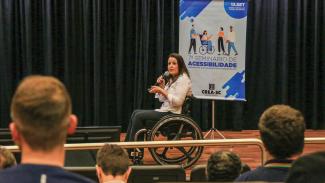 Foto da auditora fiscal de controle externo do TCE/SC Débora Borim da Silva palestrando durante o sétimo Seminário de Acessibilidade. Ela é uma mulher branca, de cabelos castanhos, cadeirante. Está no palco do auditório do TCE/SC, segurando o microfone com uma das mãos e o passador de slides com a outra. À sua frente, de costas para a foto e em primeiro plano, há pessoas que assistem à sua fala. Ao fundo, o banner do evento.