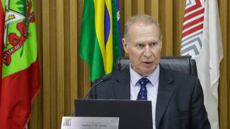 Foto do presidente do TCE/SC, conselheiro Herneus De Nadal, na sessão do Pleno. Ele está sentado e falando ao microfone. Na sua frente, um monitor de computado. Atrás dele, da esquerda para a direita, as bandeiras de Santa Catarina, do Brasil e do TCE/SC.