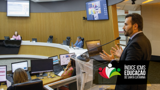 Foto mostra um homem de barba, à direita, falando ao microfone. Ele veste roupas escuras. Ao fundo, no alto, há dois televisores que projetam imagens que aparecem em computador que está em primeiro plano