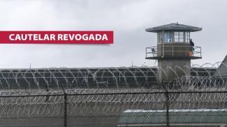 Imagem mostra o muro de uma penitenciária e uma torre de vigilância. À esquerda, escrito em branco sobre tarja vermelha, está escrito “cautelar revogada”