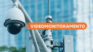 Banner horizontal com close em uma câmera de videomonitoramento instalada em poste metálico, voltada para a área externa. Ao fundo, há prédios desfocados em tons azulados. Sobre a imagem, em faixa horizontal na cor laranja, está o texto em destaque: "Videomonitoramento".