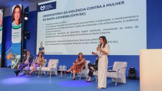 Foto da procuradora-geral Cibelly Farias palestrando no evento. Ela está em pé, falando ao microfone, no palco, que é uma estrutura azul. Atrás dela, os demais participantes, sentados em poltronas um ao lado do outro. Nas laterais, há dois telões que projetam a sua foto e nome. No telão do meio, é projetada a apresentação. À frente dos palestrantes, parte do público, de costas para a foto. 