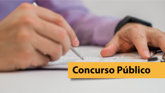 Foto que mostra uma pessoa passando a caneta sobre um papel. A foto está cortada, de modo que só é possível ver as suas mãos. Sobre a imagem, uma tarja amarela com o texto "Concurso Público". 