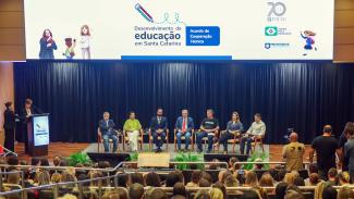 Foto horizontal dos participantes da assinatura do acordo, no palco do auditório do TCE/SC. Há sete autoridades, sentadas uma ao lado da outra. Entre os presentes, o conselheiro do TCE/SC Aderson Flores, o prefeito de Florianópolis, Topázio Neto, e a presidente executiva da Todos Pela Educação, Priscila Cruz. À frente deles, o público que assistia à solenidade, de costas, sentado em poltronas do auditório. 