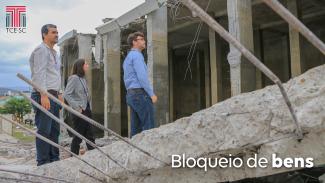 Foto dos escombros do reservatório da Casan, com colunas remanescentes, vigas caídas, com ferros expostos, e três técnicos do TCE/SC observando a área. No canto superior esquerdo há a logo do TCE/SC. E na parte de baixo, à direita, há o texto Bloqueio de bens, sobreposto a imagem, em fonte branca.