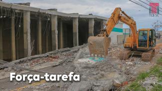 Foto dos escombros do reservatório de água da Casan, e de uma retroescavadeira fazendo a limpeza, retirando material do meio da rua. Na parte inferior direita da foto há o texto “Força-tarefa” e no canto superior direito há a logo do TCE/SC.