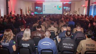 Imagem mostra em primeiro plano, e de costas, cinco profissionais da segurança sentados em meio à plateia. Nos uniformes deles, as instituições a que pertencem. Da esquerda para a direita: Polícia Científica, Polícia Civil, Bombeiros Militares, Polícia Penal e Polícia Militar. Ao fundo, participantes do seminário falam durante um dos painéis. 