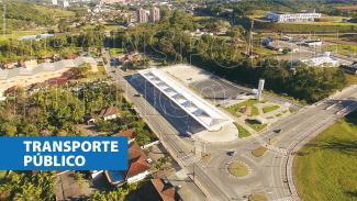 magem mostra o Terminal de Integração Norte, em Blumenau. A foto é uma panorâmica feita do alto, que mostra a estrutura utilizada pelos ônibus e um estacionamento para veículos ao lado, cercados por uma área verde 